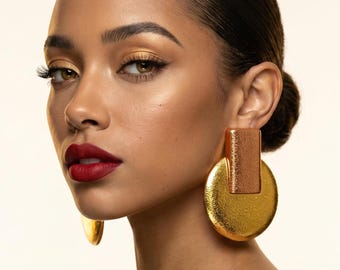 Op de afbeelding: Een vrouw met donker haar en rode lippenstift draagt grote, goudkleurige oorbellen. De oorbellen hebben een rechthoekig bovenstuk en een cirkelvormige basis. De huid van de vrouw heeft een warme tint en haar oogmake-up is goud.