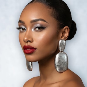 May include: A woman with dark hair styled in a bun, wearing silver textured drop earrings. Her makeup includes silver eyeshadow, black eyeliner, and red lipstick. The background is a soft, light blue.