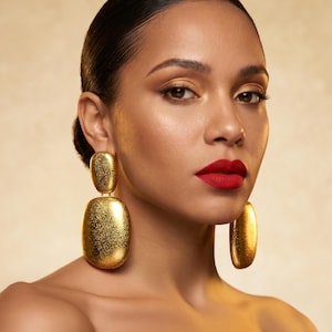 May include: A woman with dark hair and red lipstick wears large, textured gold earrings. The earrings are rectangular with rounded edges, hanging from the earlobes. The background is a warm, neutral tone, highlighting the jewelry and makeup.
