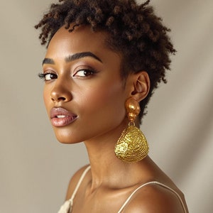 Può includere: Una donna con capelli corti e ricci castani indossa un orecchino importante dorato. L'orecchino è di forma a goccia, grande e con una texture.