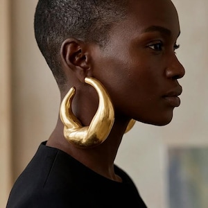 May include: A pair of large, chunky gold hoop earrings with a smooth, polished finish and a thick, sculptural, crescent-like shape. The earrings feature a rounded, organic form that tapers at the ends, creating a bold, metallic accessory.
