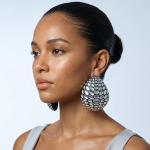 May include: Large, silver-toned teardrop earrings with a textured, honeycomb design. The earrings are worn by a person with dark hair, wearing a grey top, against a light background. The earrings are a statement piece.