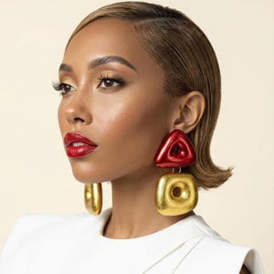 May include: A woman wearing large, geometric earrings. The earrings feature a red triangle and a gold square with a hole in the center. The woman has short, blonde hair, red lipstick, and is wearing a white top.