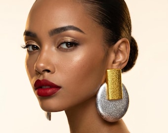 May include: Close-up of a person wearing large, statement earrings. The earrings feature a gold rectangular top and a silver, textured, circular bottom. The person has dark hair pulled back, red lipstick, and gold eyeshadow.