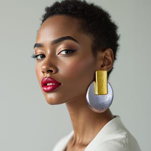 Puede incluir: Una mujer con cabello oscuro corto y maquillaje llamativo, incluyendo lápiz labial rojo y delineador de ojos alado, lleva pendientes grandes y llamativos. Los pendientes presentan una parte superior rectangular dorada y una base circular plateada brillante.