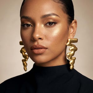 May include: A woman with gold statement earrings. The earrings are a flowing, abstract design in gold. She is wearing a black top. The background is a neutral beige colour.