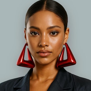 May include: A close-up shot of a person wearing large, shiny, red triangle-shaped earrings. The earrings have a hollowed-out design and are a bold statement piece. The person is wearing a dark blazer against a light blue background.