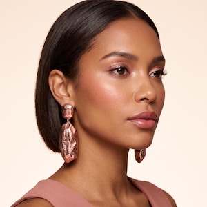 May include: Rose gold statement earrings with a textured, organic design. The earrings are worn by a person with dark hair and a pink top. The earrings have a rectangular top and a larger, irregular shape hanging below.