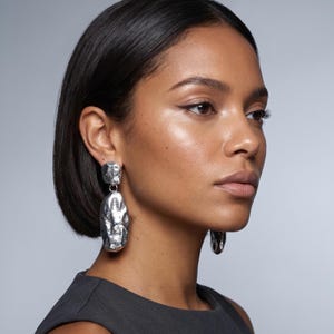 May include: Silver-toned, textured drop earrings. The earrings feature a square top and an elongated, organic shape below. The jewelry is worn by a person with dark hair and a gray top. The background is a solid gray.