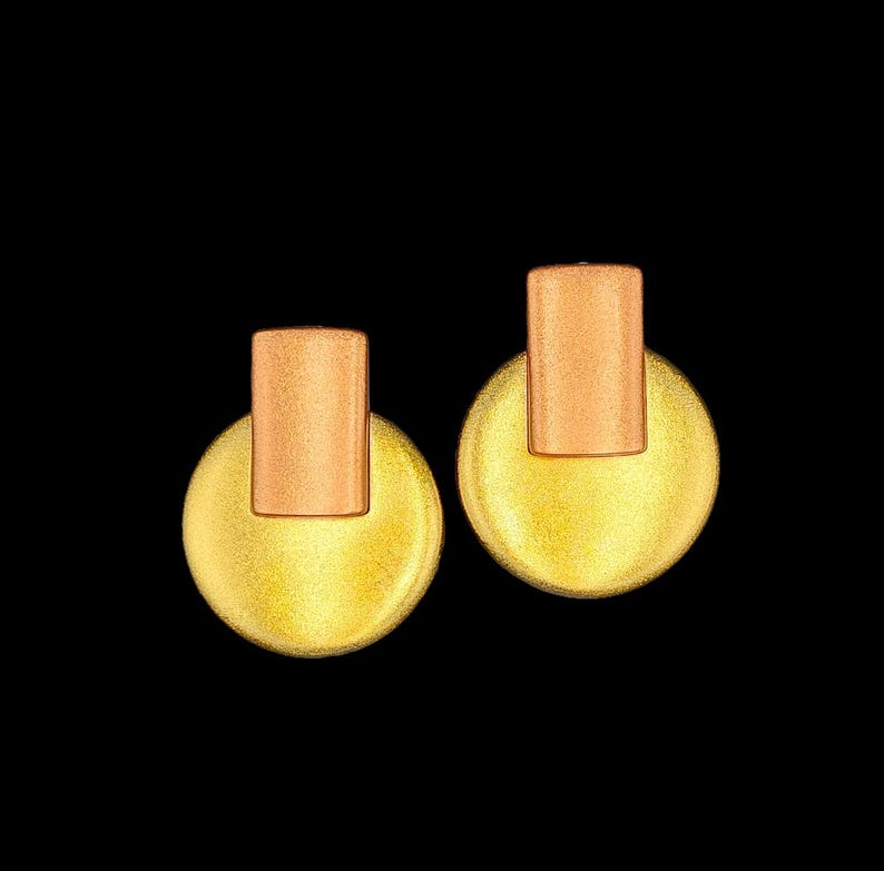 May include: A pair of gold-coloured stud earrings. Each earring has a circular, textured base with a rectangular top piece. The earrings have a warm, metallic finish, set against a black background.