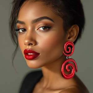 Może przedstawiać: Kobieta o ciemnych włosach i czerwonej szmince nosi duże, czerwone, spiralne kolczyki. Kolczyki są wykonane z miękkiego, przypominającego tkaninę materiału.