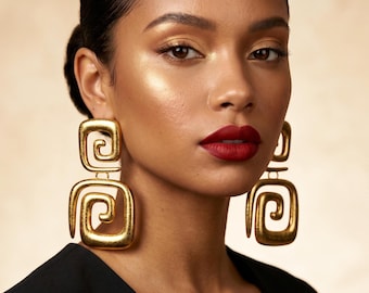 Può includere: Una donna indossa grandi orecchini geometrici color oro. Gli orecchini hanno un design a doppio quadrato con un motivo a spirale. Indossa un top nero e rossetto rosso. Lo sfondo è di un colore neutro e morbido.