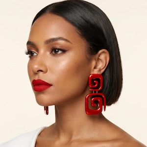 May include: Bold, red, geometric earrings with a square and spiral design. The earrings are a vibrant, glossy red and are worn by a person with dark hair and red lipstick. The earrings are a statement piece.