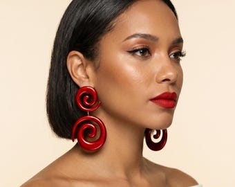 May include: A woman wearing large, red, spiral-shaped earrings. The earrings are a bold, glossy red and hang from the earlobes. The woman has dark hair, red lipstick, and is wearing a white top. The background is a neutral beige.