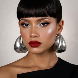 May include: A woman with dark hair and bold makeup, including silver eyeshadow and deep red lipstick, is wearing large, silver hoop earrings. The earrings are a prominent feature, reflecting light. The woman is wearing a black top.