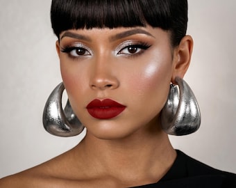 May include: A woman with dark hair and bold makeup, including silver eyeshadow and deep red lipstick, is wearing large, silver hoop earrings. The earrings are a prominent feature, reflecting light. The woman is wearing a black top.