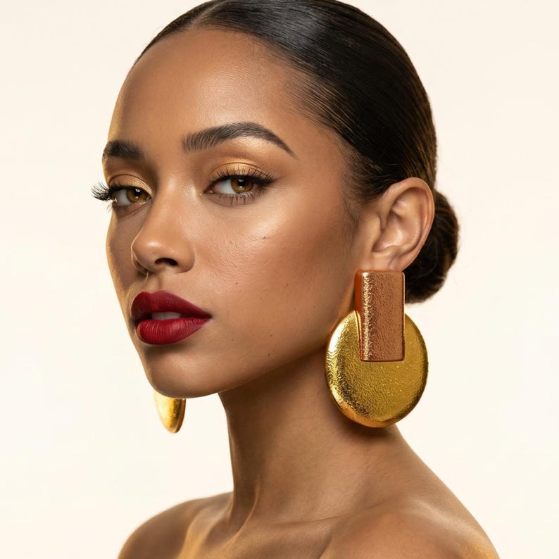 May include: A woman with dark hair and red lipstick is wearing large, gold earrings. The earrings have a rectangular top and a circular base. The woman's skin has a warm tone, and her eye makeup is gold.