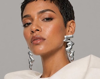 Può includere: Una persona con capelli corti e scuri indossa grandi orecchini astratti color argento. Gli orecchini hanno una forma fluida e irregolare. Indossa un top color crema su uno sfondo grigio.