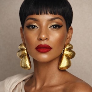Puede incluir: Primer plano de una persona con pendientes dorados grandes y texturizados. Los pendientes tienen un diseño abstracto único. La persona tiene el pelo corto y oscuro, sombra de ojos dorada y lápiz labial rojo. El fondo es beige neutro.