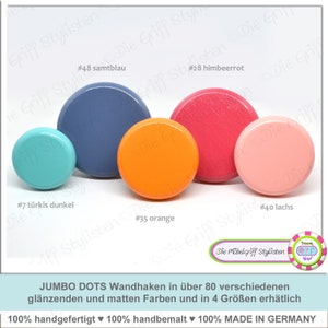 Könnte beinhalten: Vier Holz-Knöpfe in verschiedenen Farben: Türkis, Blau, Orange und Rosa. Die Knöpfe sind mit Nummern und Farbnamen beschriftet. Der Text darunter lautet: "JUMBO DOTS Wandhaken in über 80 verschiedenen glänzenden und matten Farben und in 4 Größen erhätlich 100% handgefertigt ♥ 100% handbemalt ♥ 100% MADE IN GERMANY".