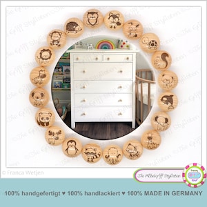 May include: A collection of wooden cabinet knobs with engraved animal designs, including a lion, zebra, and koala. The knobs are arranged around a white dresser. The image also includes text that says "Die Möbelgriff Stylisten" and "MADE IN GERMANY".