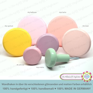 May include: Assortment of round and knob-shaped wooden wall hooks in various pastel colors, including pink, yellow, green, and lavender. The hooks are labeled with numbers and color names in German. The text at the bottom reads "Wandhaken in über 80 verschiedenen glänzenden und matten Farben erhältlich 100% handgefertigt ♥ 100% handbemalt ♥ 100% MADE IN GERMANY".