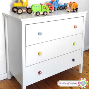 Puede incluir: Una cómoda blanca con cuatro cajones y tiradores de cajones en forma de coches de juguete de colores. La cómoda tiene una tapa de madera clara y está sentada en un suelo de madera. El texto "Die Möbelgriff Stylisten" es visible en el suelo frente a la cómoda.
