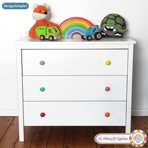 May include: White dresser with three drawers and colorful knobs. The knobs are purple, green, red, yellow, and pink. The dresser is against a white wall with a rainbow arch, a toy truck, a toy car, a stuffed fox, and a stuffed turtle on top.