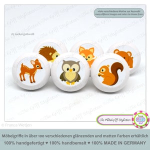 以下が含まれることがあります： カラフルな動物のイラストが描かれた白い木製ノブ6個セット。動物は鹿、フクロウ、ハリネズミ、キツネ、リス、クマ。ノブには「#3 zuckergußweiß」と「Die Möbelgriff Stylisten」のラベルが貼られています。