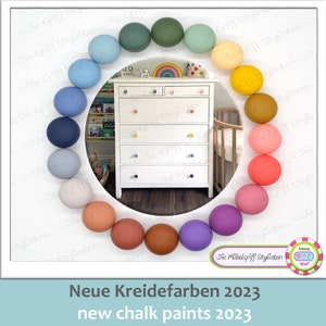Może przedstawiać: Koło kolorowych drewnianych kulek otacza białą komodę z sześcioma szufladami. Komoda ma kolorowe gałki. Tekst "Neue Kreidefarben 2023" i "new chalk paints 2023" znajduje się na dole obrazu.