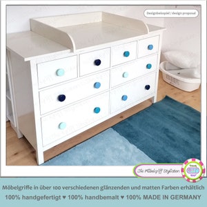 Peut inclure: Une commode blanche avec une table à langer sur le dessus. La commode a neuf tiroirs avec des boutons bleus et turquoise. La commode est dans une pièce avec un tapis bleu clair et bleu foncé.