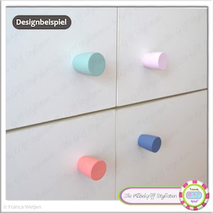 Peut inclure: Un meuble à quatre tiroirs avec des boutons de différentes couleurs. Les boutons sont bleu clair, rose, bleu et rose clair. Le meuble est blanc et porte l'inscription "Designbeispiel" en haut.