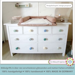 Peut inclure: Une commode blanche à six tiroirs avec des boutons de couleur pastel. La commode est surmontée d'un matelas à langer et d'un tapis à langer blanc. Le texte "Designbeisspiel / design proposal" est dans le coin supérieur droit de l'image. Le texte "Die Möbelgriff Stylisten" est dans le coin inférieur droit de l'image. Le texte "Möbelgriffe in über 100 verschiedenen glänzenden und matten Farben erhältlich 100% handgefertigt ♥ 100% handbemalt ♥ 100% MADE IN GERMANY" est en bas de l'image.