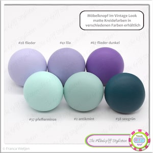 May include: Assortment of six round wooden knobs in various colors, including lavender, light blue, teal, and mint green. The knobs are labeled with numbers and color names, such as #28 flieder, #41 lila, #67 flieder dunkel, #37 pfefferminze, #2 antikmin, and #38 seegrün. The text "Möbelknopf im Vintage Look matte Kreidefarben in verschiedenen Farben erhältlich" is also visible in the image.