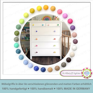 Può includere: Una cassettiera bianca con sei cassetti, ognuno con una manopola colorata. Le manopole sono disposte a forma di arcobaleno. La cassettiera è circondata da un cerchio di manopole colorate in varie tonalità, tra cui verde, blu, giallo, rosa e marrone.