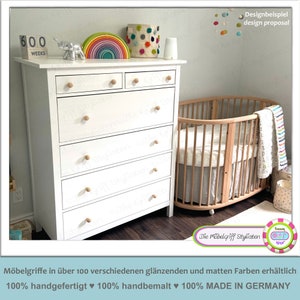 Peut inclure: Une commode blanche à six tiroirs avec des boutons en bois. La commode est dans une chambre d'enfant avec un berceau en bois et un jouet arc-en-ciel coloré. Le texte "600 WEEKS" est sur un bloc de bois sur le dessus de la commode.