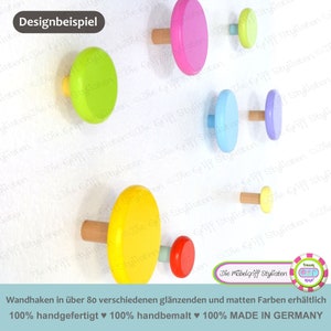May include: Colorful wooden wall hooks in various shapes and sizes, including yellow, green, pink, blue, and red. The hooks are hand-painted and made in Germany. The text on the image reads "Wandhaken in über 80 verschiedenen glänzenden und matten Farben erhältlich 100% handgefertigt ♥ 100% handbemalt ♥ 100% MADE IN GERMANY"