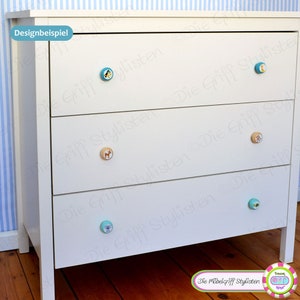 May include: White dresser with three drawers and decorative animal knobs. The knobs are blue, brown, and beige. The dresser is against a blue and white striped wall.