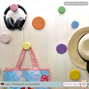 Op de afbeelding: Een verzameling kleurrijke, ronde wandhaken in verschillende tinten, waaronder roze, oranje, groen, blauw, paars en geel. Een zwarte koptelefoon, een draagtas en een strohoed hangen aan de haken. De haken zijn bevestigd op een witte houten muur.