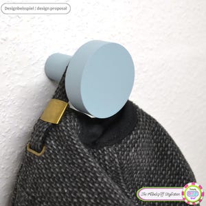 Peut inclure: Un crochet mural en bois bleu clair de forme circulaire. Un manteau gris foncé avec une boucle dorée est suspendu au crochet. Le texte "Designbeispiel / design proposal" est dans le coin supérieur gauche.
