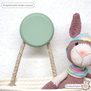 Peut inclure: Un crochet mural rond en bois vert clair, soutenu par deux cordons en corde à motifs. Un lapin crocheté portant un chapeau et une écharpe est en arrière-plan. Le texte "Designbeispiel / design proposal" est en haut.