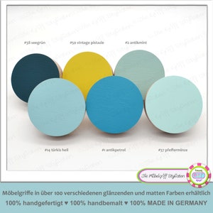Può includere: Cinque pomelli rotondi in legno in varie tonalità di blu e verde. I pomelli sono etichettati con numeri e nomi di colori, tra cui verde mare, pistacchio vintage, menta antica, turchese chiaro e menta piperita. Il testo "Die Möbelgriff Stylisten" è visibile nella parte inferiore dell'immagine.