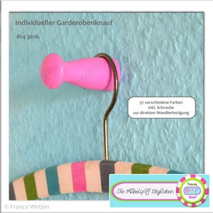 Puede incluir: Pomo de madera rosa con un gancho dorado, etiquetado '#14 rosa' y 'Individueller Garderobenknauf'. El pomo está fijado a una pared azul texturizada. El texto '57 verschiedene Farben inkl. Schraube zur direkten Wandbefestigung' es visible en la imagen.
