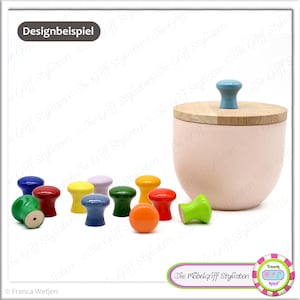 以下が含まれることがあります： カラフルな木製ノブのコレクションと、木製の蓋と青いノブが付いたピンク色の容器。ノブは青、赤、黄、緑、オレンジなど様々な色があります。「Die Möbelgriff Stylisten」の文字が見えます。
