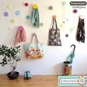 Könnte beinhalten: Eine weiße Wand mit bunten runden Haken, an denen einige Schals hängen. An den Haken hängen zwei Stofftaschen mit Blumenmuster, eine gelbe und eine rosafarbene. Ein schwarzer Regenschirm steht in einem Metall-Regenschirmständer. Der Text auf dem Ständer lautet "Die Möbelgriff Stylisten" und "Troom Ring".