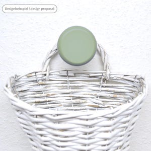 May include: A white wicker basket with a distressed finish, hanging from a light green circular hook. The basket has a woven design and a handle. The text "Designbeispiel / design proposal" is in the upper left corner.