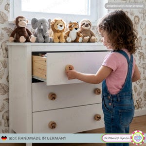 Può includere: Una cassettiera in legno bianco con tre cassetti dotati di pomelli rotondi in legno chiaro. Ogni pomello è inciso con una piccola illustrazione di un animale, tra cui una scimmia, un leone e una tigre. Il cassetto superiore è parzialmente aperto.