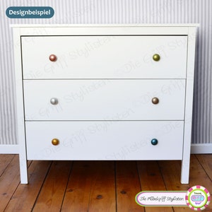 May include: White dresser with three drawers and four different colored round knobs. The dresser is on a wooden floor.