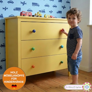 Könnte beinhalten: Eine hellgelbe Holzkommode mit drei Schubladen und bunten, runden Knöpfen in Blau, Rot, Grün und Orange. Die Kommode steht vor einer hellblauen Wand mit einem sich wiederholenden Muster aus dunkelblauen Fahrzeugen und Flugzeugen.
