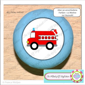以下が含まれることがあります： 赤い消防車のイラストが描かれた青い丸いノブ。ノブには「#23 blau mittel」と「Die Möbelgriff Stylisten」のラベルが貼られています。